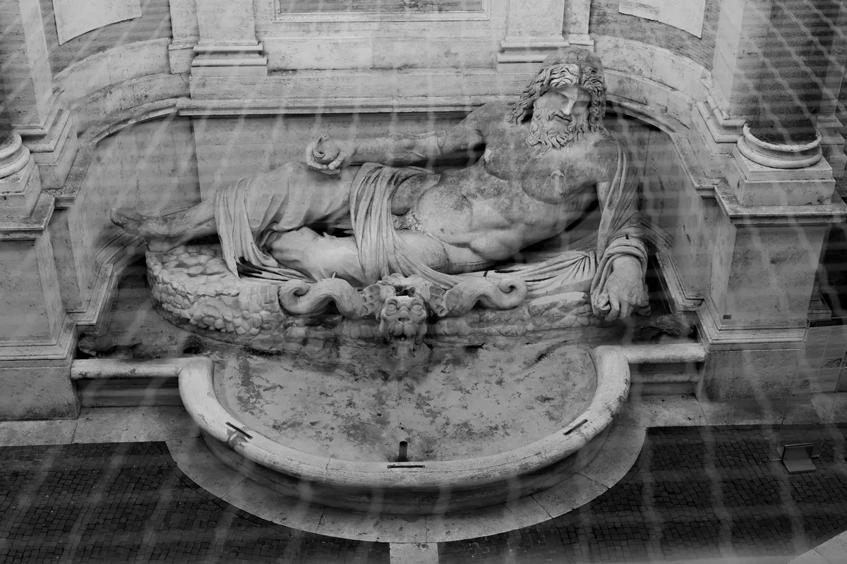 Black and white photo of an ancient marble statue resembling a reclining Greek or Roman god in an ornate fountain.